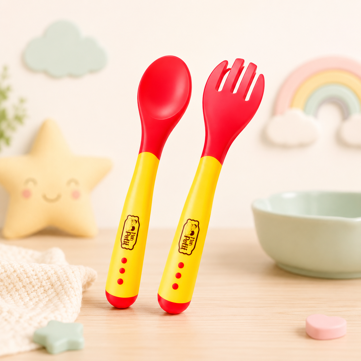 Conjunto de colher e garfo Mon Petit colocado na vertical sobre uma mesa num cenário infantil decorado com uma almofada de estrela sorridente, um arco-íris pastel e uma tigela verde.