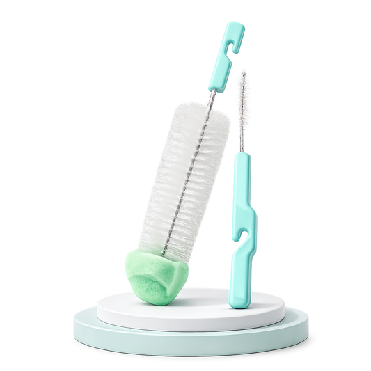 Kit de limpeza infantil contendo uma escova grande com ponta de esponja verde-clara e uma escova fina para bicos, ambas com cabos verde-água, expostas num pódio circular sobre fundo branco.